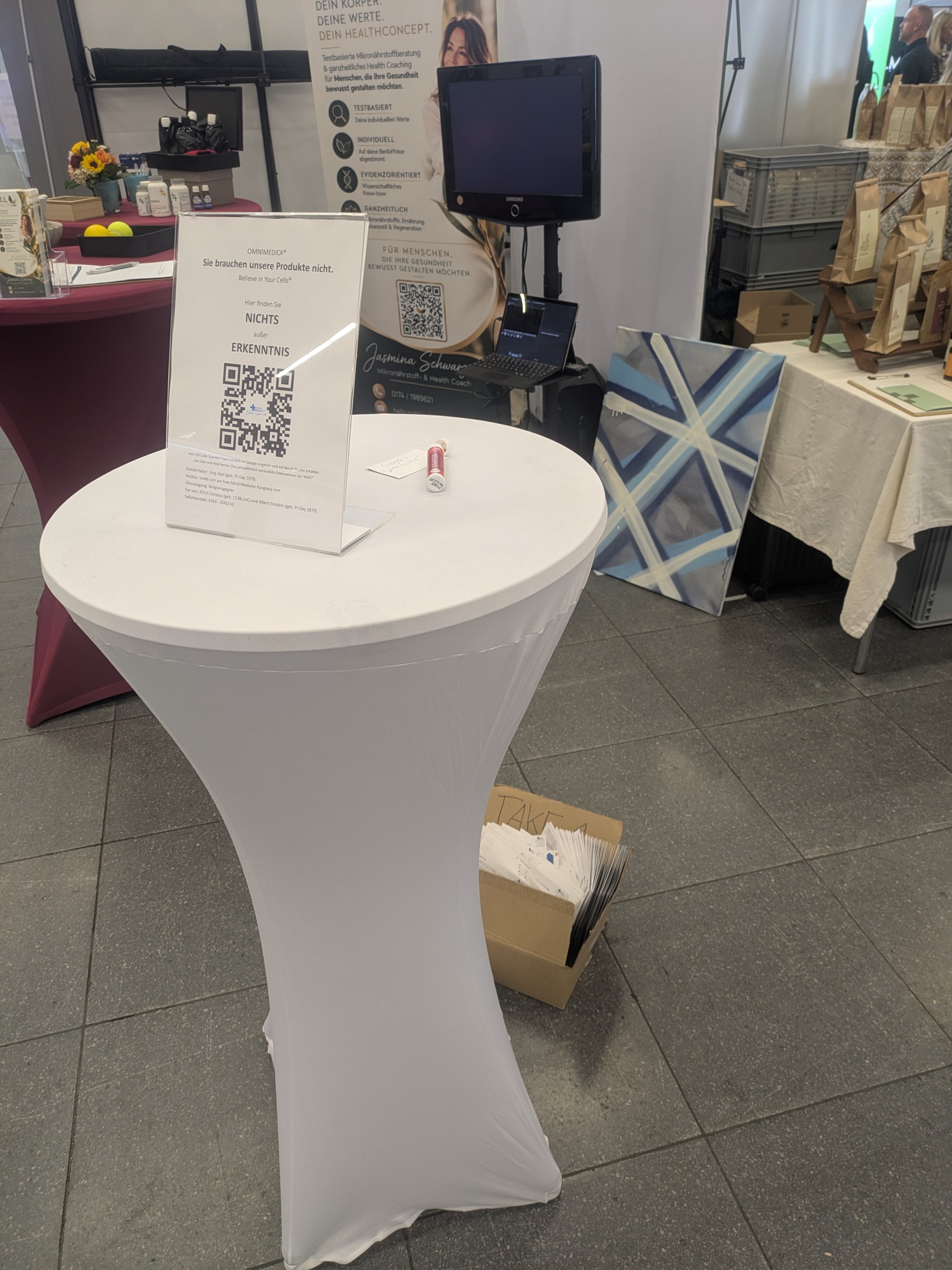 OMNIMEDICA-Stand auf dem Free Mind Medicine Kongress 2026 mit Stehtisch, Schild ‚Sie brauchen unsere Produkte nicht‘, QR-Code und einem einzelnen OM24 Starter.
